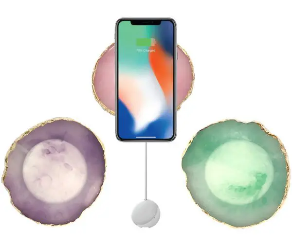 Smartphone with charging indicator on screen, flanked by three geode slices in purple, pink, and green, and a white charging cable connected to the phone.