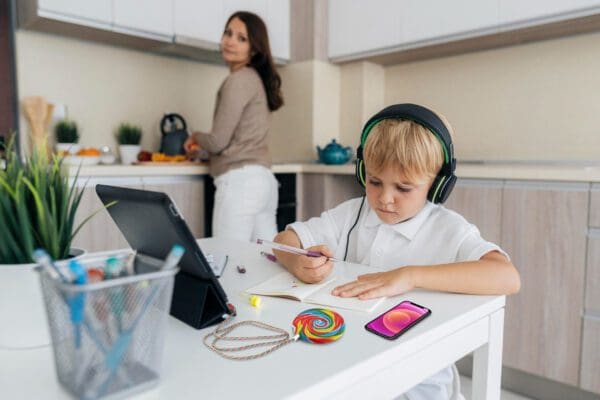 A young boy wearing headphones uses a tablet at a kitchen table while a woman works in the background.