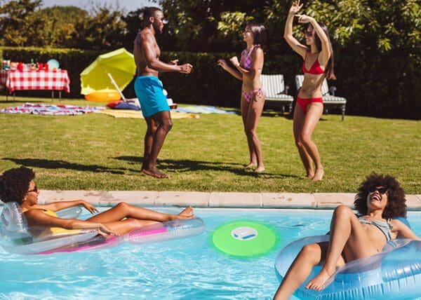 Group of friends enjoying a sunny day at a pool party, with two people playing with a beach ball and two others floating in the pool on inflatable rings.