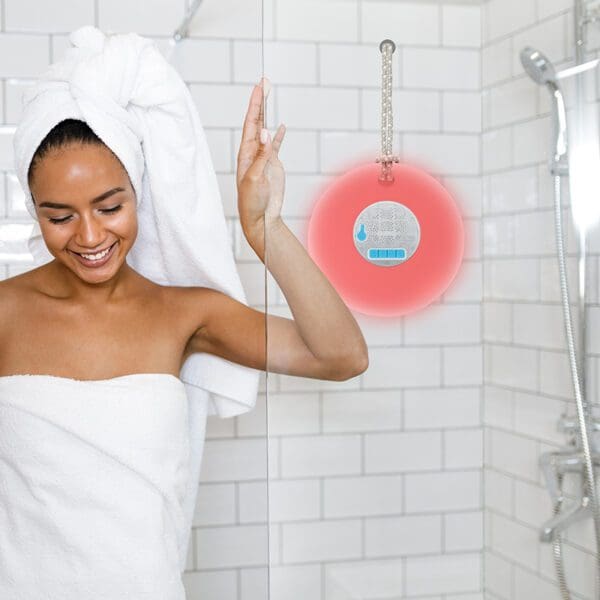 A smiling woman in a towel and head wrap reaches for a waterproof speaker in a shower.