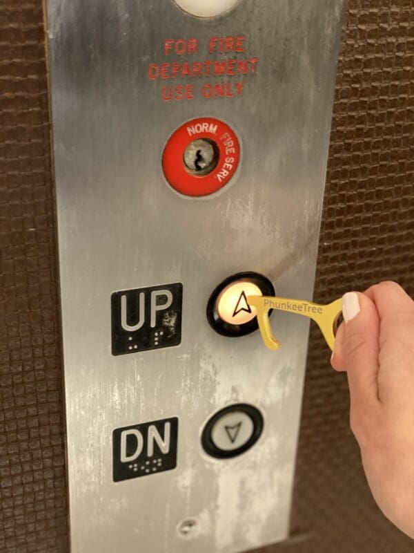 A hand pressing the 'up' button on an elevator control panel with labeled buttons for directions and emergency use.
