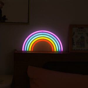 A colorful neon rainbow nightlight illuminates a dark room, placed on a wooden headboard.