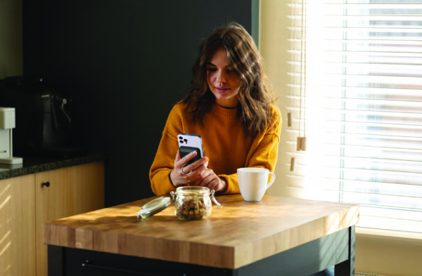 A woman in a yellow sweater smiles at her phone while standing in a kitchen with a coffee mug and a jar of cookies nearby.