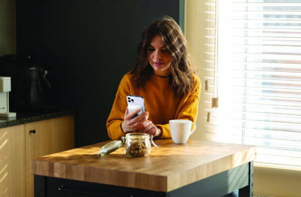 A woman in a yellow sweater smiles at her smartphone in a kitchen, with a coffee cup and a jar of cookies nearby.