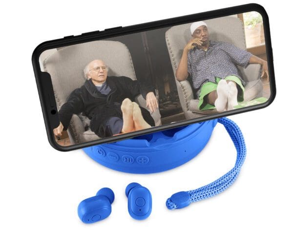 Smartphone on a blue stand displaying a video call between two elderly men, one in an armchair, accompanied by wireless earbuds.