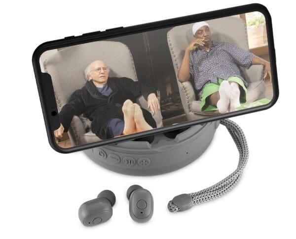 Two elderly men, one holding his head in thought and the other looking away, appear on a smartphone screen, placed on a wireless charger with earbuds beside it.