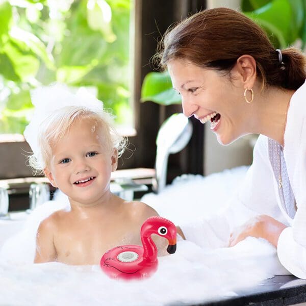 A joyful woman and a toddler with a bubble bath hat laugh together beside a toy flamingo float in a bathtub.
