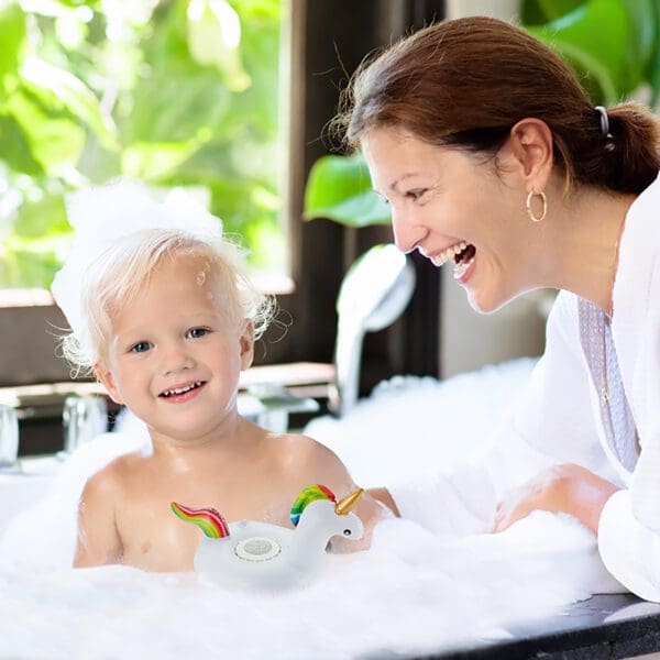 A woman and a toddler smiling and playing with a rubber duck in a bubble bath.