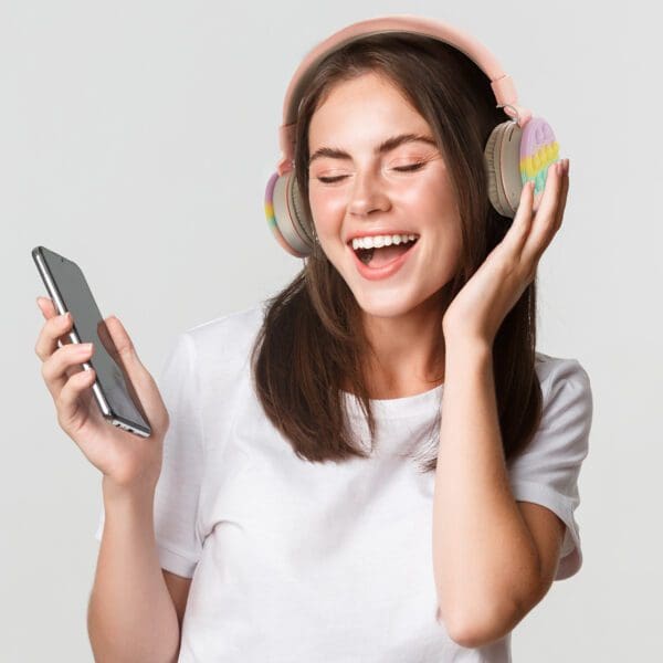 A joyful woman wearing headphones and holding a smartphone, laughing and looking at the device, on a light gray background.