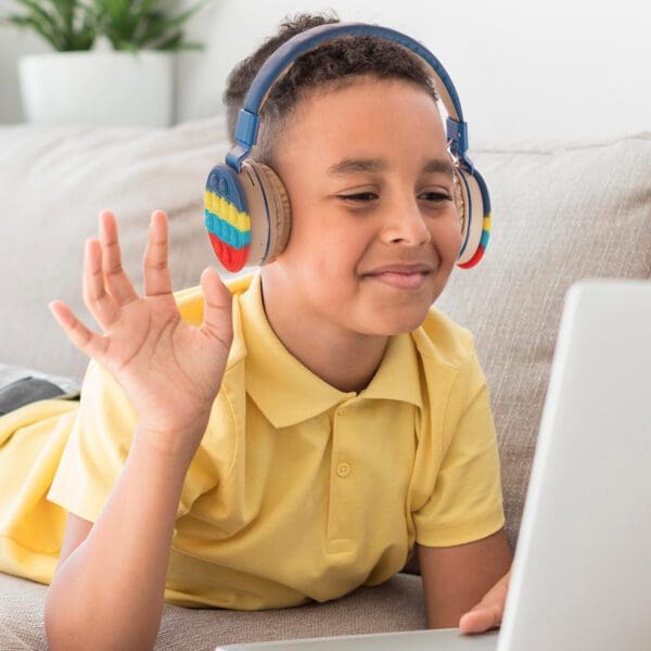 Young boy in a yellow shirt, wearing headphones, waves at a laptop screen while smiling, sitting indoors.