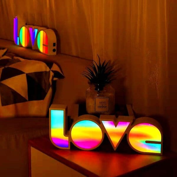 Illuminated "love" sign in rainbow colors on a shelf, with a decorative pineapple and other objects in a dimly lit room.