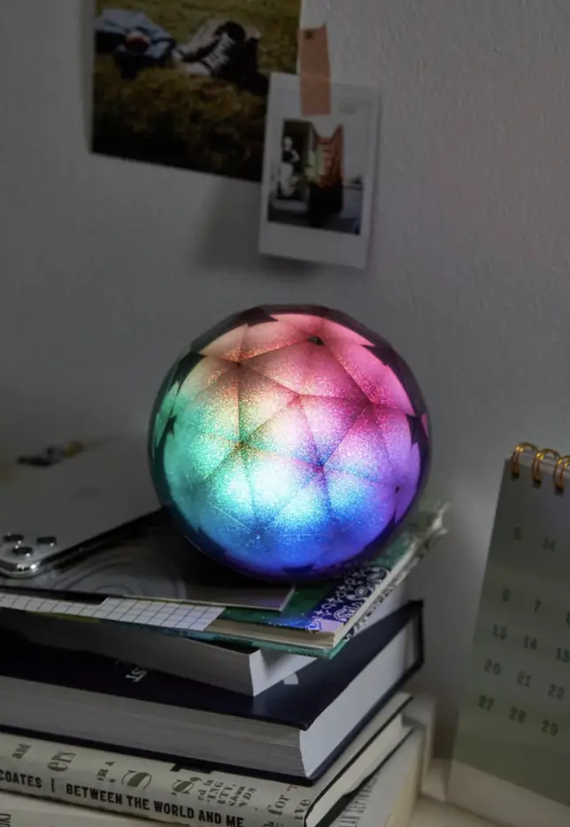 A colorful LED Crystal Ball Wireless Speaker on a stack of books beside a calendar and wall with photos and notes.