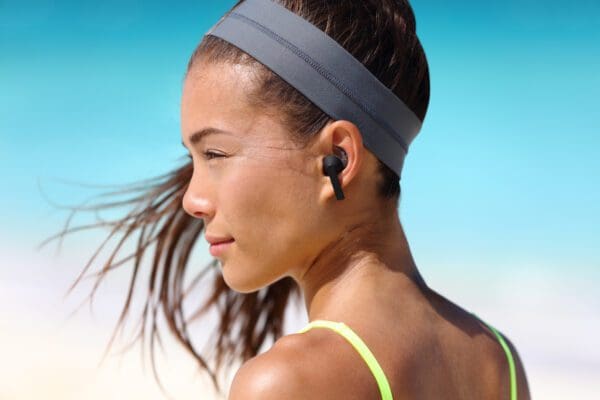 Profile view of a young woman wearing Solar Wireless Earbuds and a headband against a clear blue sky, with her hair blowing in the wind.