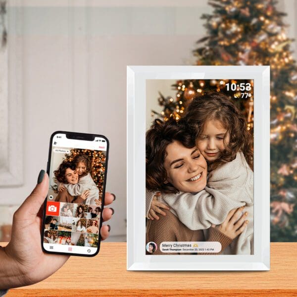 Sentence with replaced product name: A person's hand holding a smartphone displaying a photo of a smiling woman and child hugging, matched by a Digital Picture & Video Frame with an app showing the same image beside a Christmas tree.