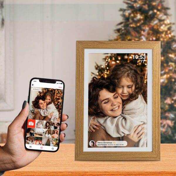 A person's hand holding a smartphone displaying a photo of a mother and daughter embracing, with a Digital Picture & Video Frame showing the same image nearby; a Christmas tree is in the background.