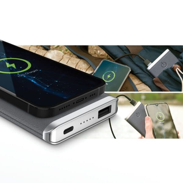 Two smartphones charging on portable power banks; one via cable and the other on a Grey - Leather 5K Wireless Charging Power Bank, displayed on a table with a backpack in the background.