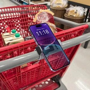 A smartphone displaying the time "11:18" is placed on a red Phone Mount Clamp attached to a shopping cart handle in a supermarket aisle.